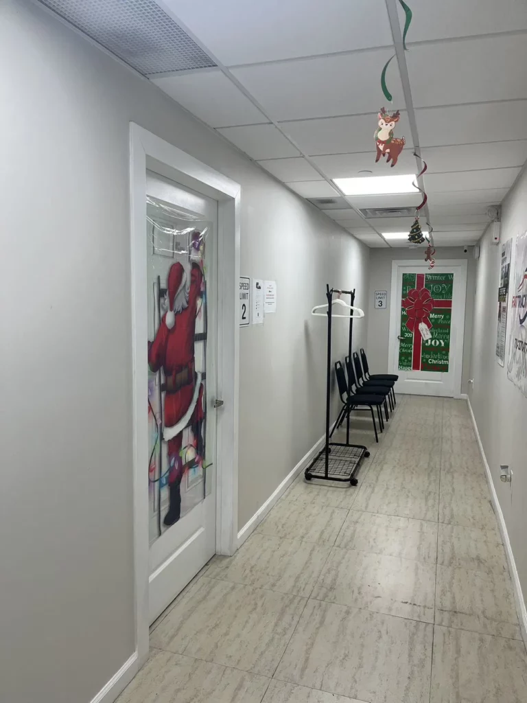 A hallway with white walls and a light-colored tiled floor. Decorations include a Santa Claus figure on a door and hanging ornaments from the ceiling. Several black chairs and a coat rack are along the wall, and the far door has festive holiday-themed wrapping.