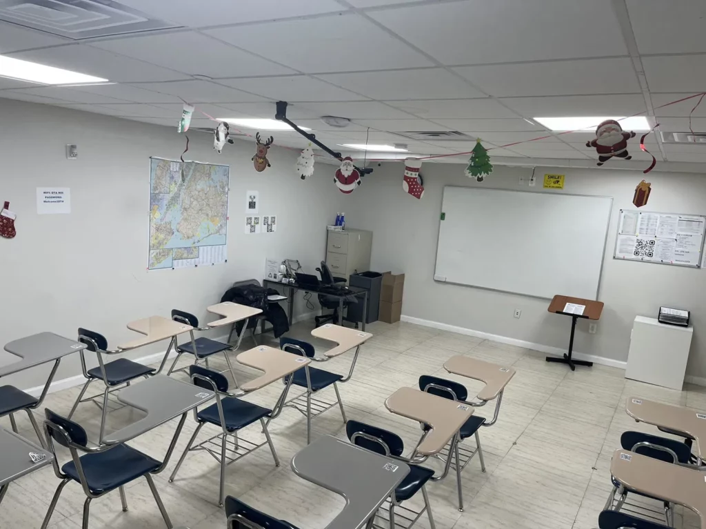 A classroom with light-colored walls is decorated with holiday ornaments hanging from the ceiling. There are multiple desks arranged in rows facing a whiteboard at the front. A map is displayed on the wall, and several documents and decorations are visible around the room.