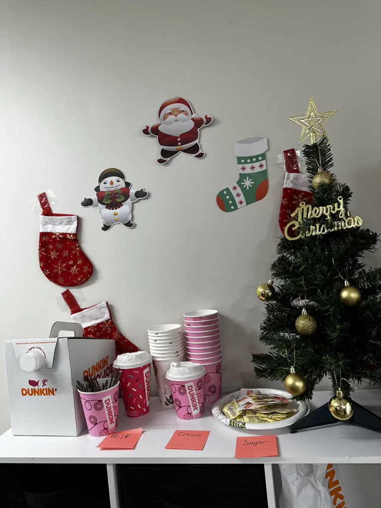 A festive setup features a small Christmas tree decorated with gold ornaments and a topper reading "Merry Christmas." Nearby, Dunkin' cups, bags, napkins, and containers are arranged on a table. Decor includes stockings, a Santa, and a penguin cut out on the wall.
