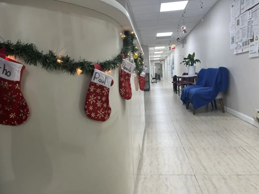 A hallway is decorated for Christmas with four red stockings labeled with names and hung on a white curved counter adorned with garland and fairy lights. The hallway has blue chairs covered with cloths, a plant, and various posters on the wall.