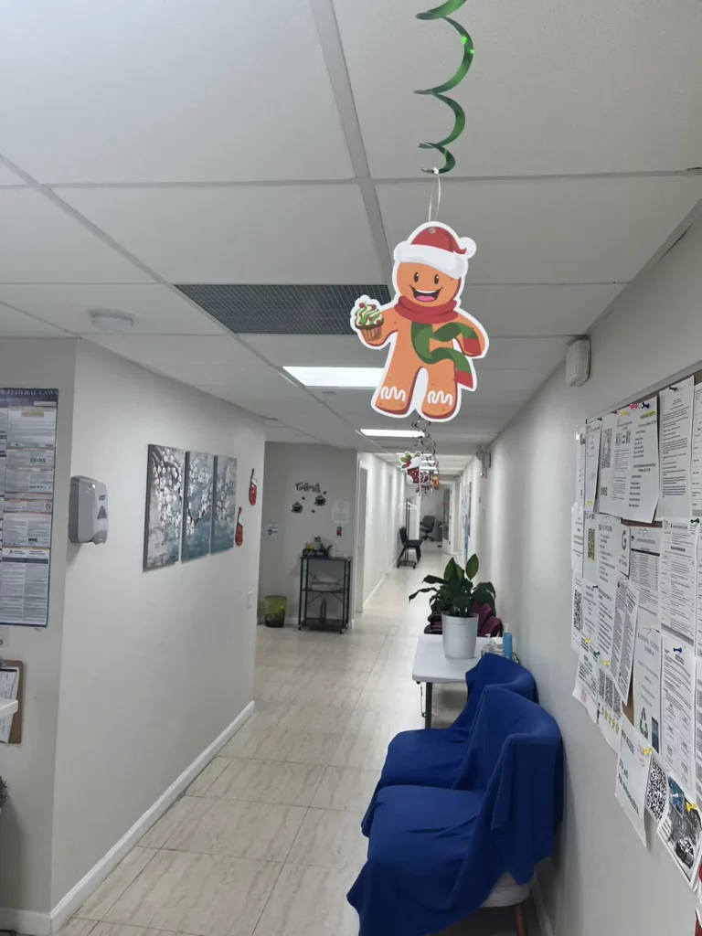 A hallway decorated for the holidays. Hanging from the ceiling are gingerbread man decorations wearing Santa hats. The walls are adorned with various posters and flyers. A blue draped bench sits against the wall with a potted plant on it.