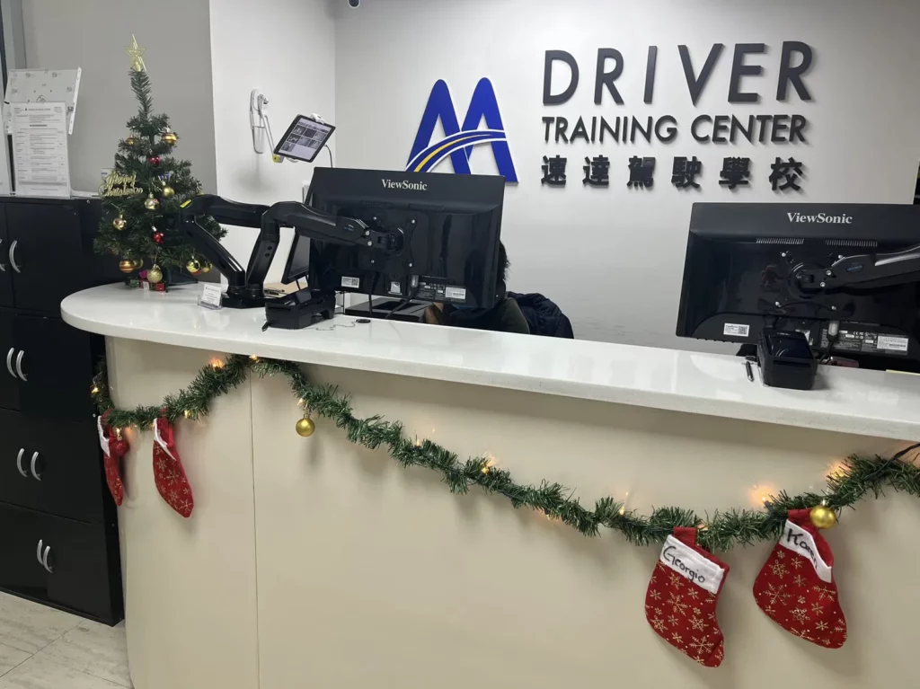Reception desk of a driver training center decorated with Christmas ornaments, including stockings, a garland, and a small Christmas tree. Two computer monitors are on the desk, and signage in English and Chinese is visible on the wall behind.