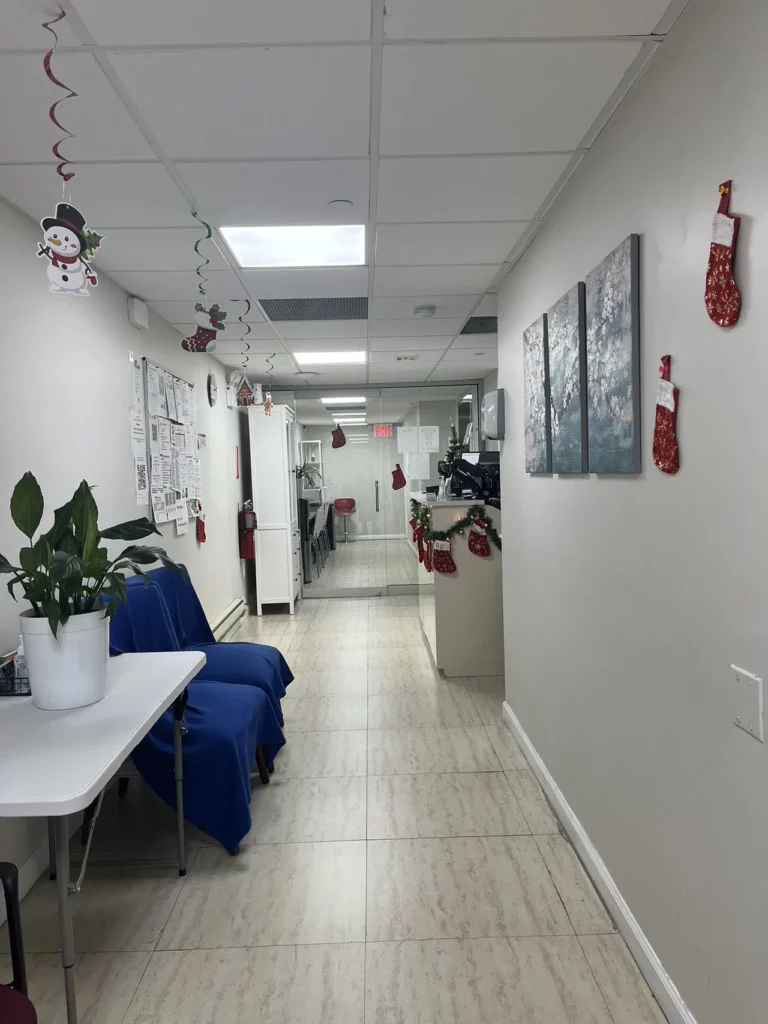 A bright corridor with holiday decorations including red stockings and hanging snowmen. Blue-covered chairs sit beside a table with a potted plant on the left. Bulletin boards and artworks adorn the white walls. Further down, there are desks and a red exit sign.
