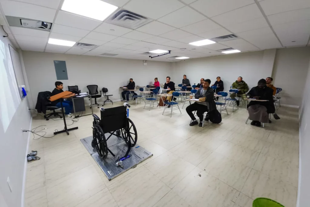 A classroom with white walls and tile floors has students sitting at individual desks. A person is speaking behind a podium at the front, next to projection screens. A wheelchair is placed on a platform at the front. The students are attentively listening.