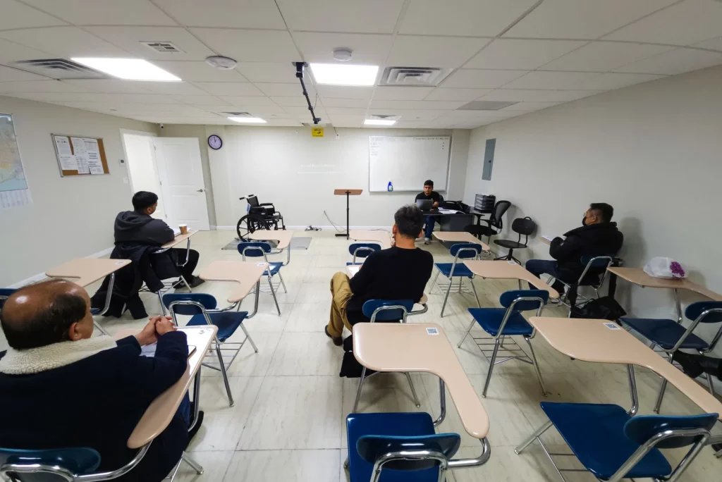 A classroom with five men seated at desks facing a podium and whiteboard at the front. One man uses a wheelchair. The room has beige walls, a clock, and fluorescent lighting. The desks are arranged in rows, and some chairs are empty.
