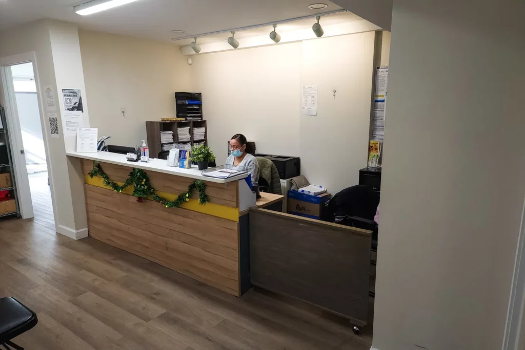 A reception area in an office, featuring a receptionist seated behind a desk decorated with a green garland for a festive touch. The receptionist is wearing a mask and working at a computer, with various office supplies neatly organized on the desk. The space is clean and well-lit, with a wooden floor and a professional yet welcoming atmosphere. Shelves behind the desk hold stacks of papers and additional office supplies
