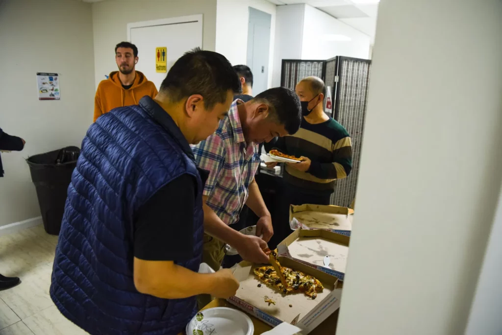 A group of people are standing around an open pizza box inside a room. Two individuals in the foreground are picking up pizza slices, while others in the background are conversing. The setting appears to be casual and indoor.
