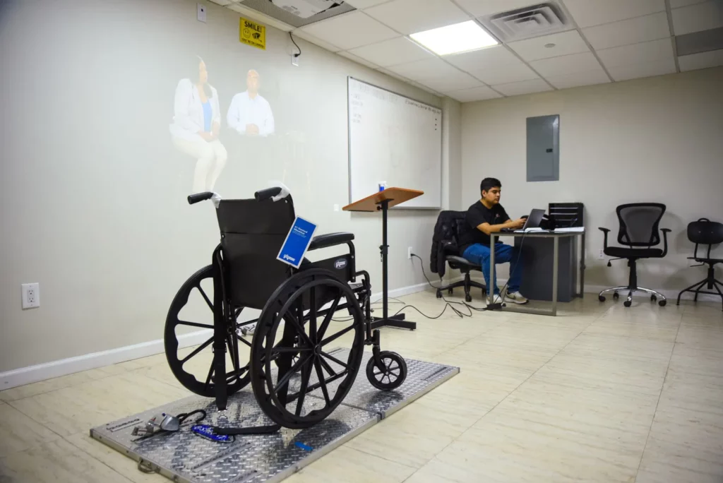 A room with beige walls and tiled flooring contains a black manual wheelchair on a metal platform. In the background, a man works on a laptop at a desk. An overhead projection screen displays an image of two people. Several empty chairs are also present.