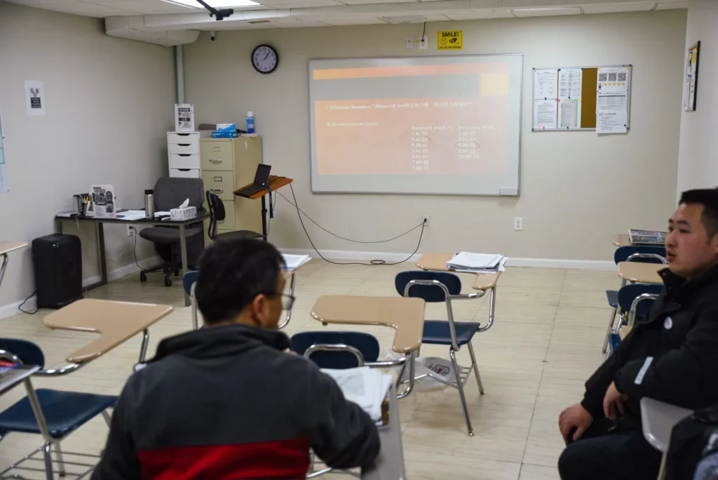 A classroom with several desks, two men seated, one in a red and black jacket looking at the other, who is in a black coat. The wall has a projection screen displaying a presentation. Chairs and tables are arranged in rows, and a clock shows the time as 4:14.