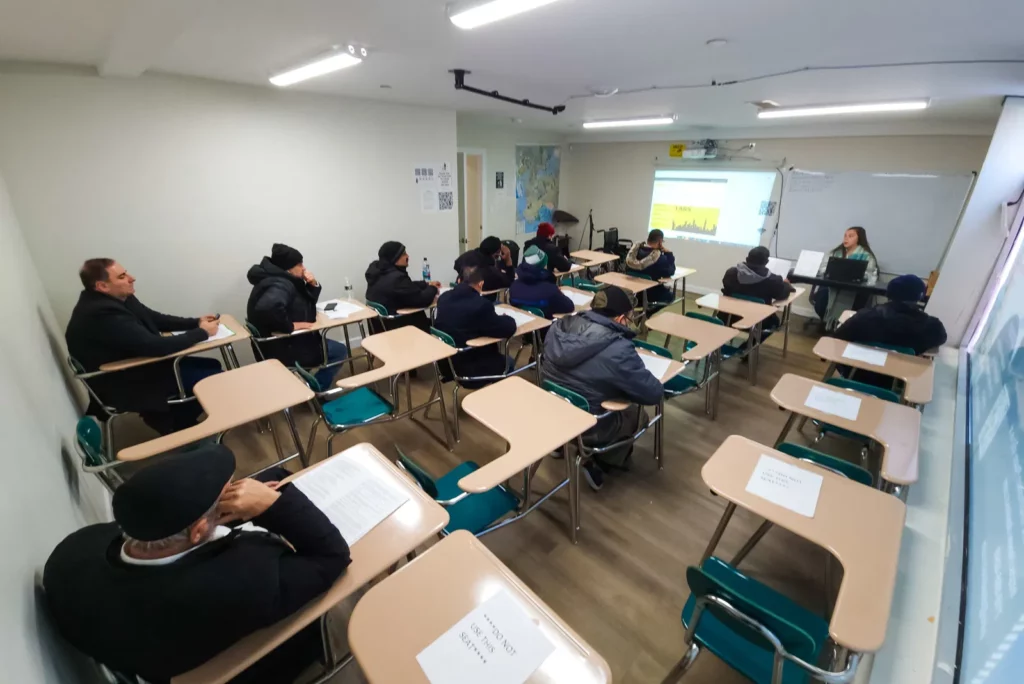 A classroom with students seated at individual desks, facing a projector screen at the front of the room where an instructor is teaching. The students are wearing winter clothing, indicating it may be cold outside. The projector displays an image related to the lesson, and there is a map on the wall. The environment is focused and conducive to learning, with students engaged in the material presented