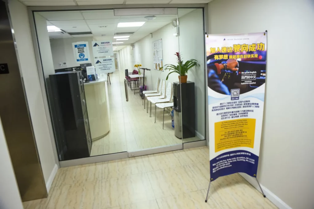 A clean, modern office lobby with white walls and floor. It features a row of white chairs against one wall and a potted plant beside the glass entrance doors. A colorful standing banner with text in Chinese is placed near the entrance.