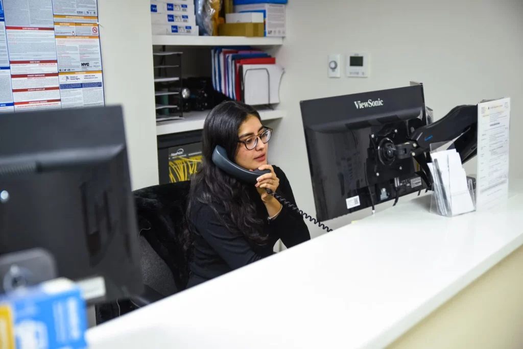 A woman with long dark hair and glasses sits at a desk in an office, speaking on the phone. She is in front of a computer monitor, with shelves and office supplies in the background. She appears focused and engaged in the conversation.