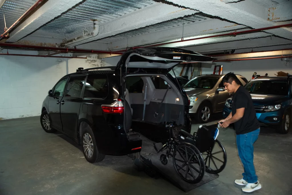 A person in blue pants and a black shirt is using a ramp to load a wheelchair into the back of a black van in a parking garage. Other vehicles are parked in the background.