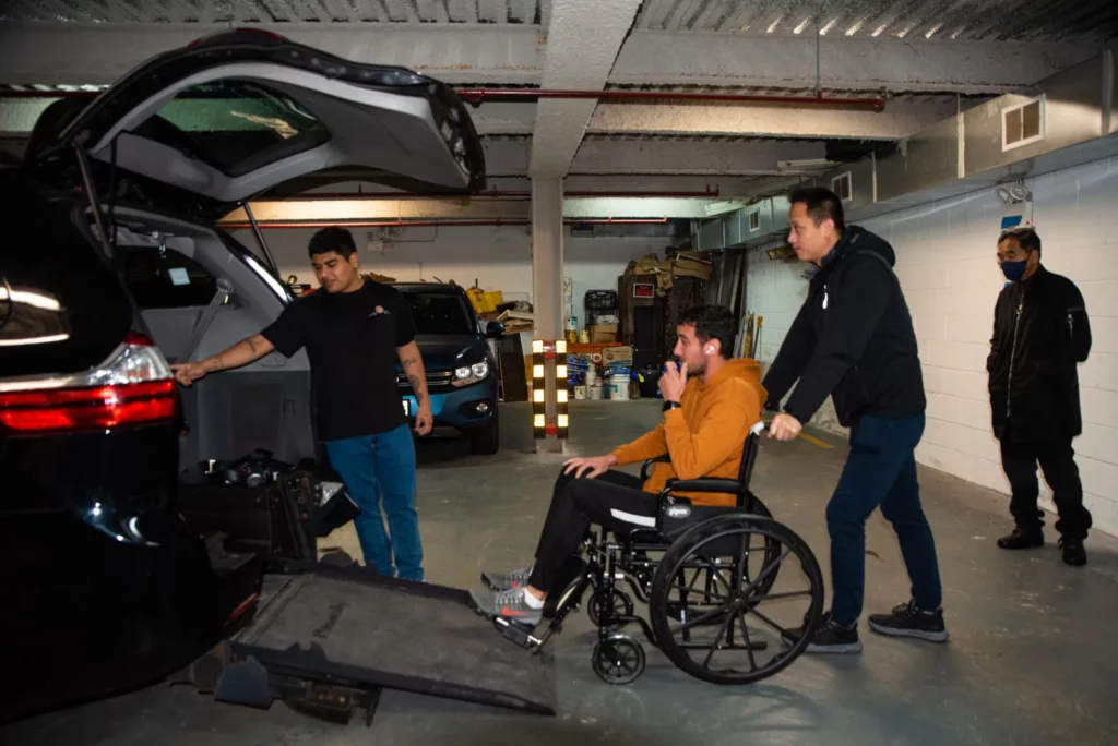 A person in a wheelchair is being assisted by two individuals to enter a vehicle via a ramp. Another person stands by the car, and a fourth person watches from a distance. They are in an indoor parking area with boxes and equipment in the background.