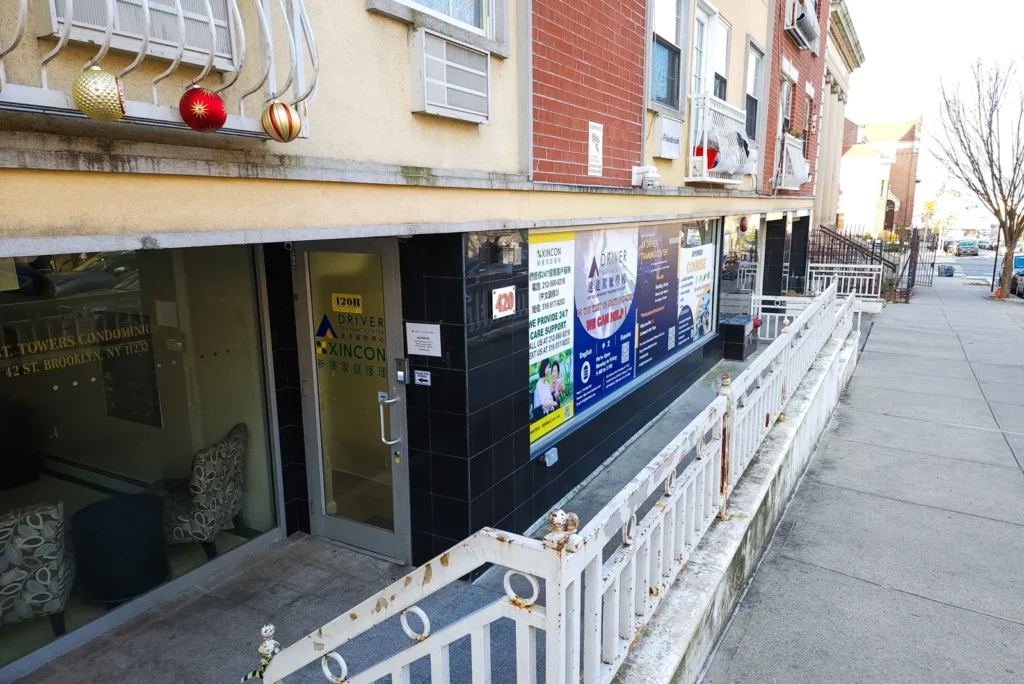 An exterior view of a building entrance, featuring a glass door labeled with signage for ‘Xincon Driver Training Center’ and ‘Towers Condominium’ located at 142 St. Brooklyn, NY. The sidewalk and exterior railing lead up to the entrance, with holiday ornaments hanging from the balcony above. The building has a clean and professional appearance, with various informational posters displayed on the windows along the sidewalk.