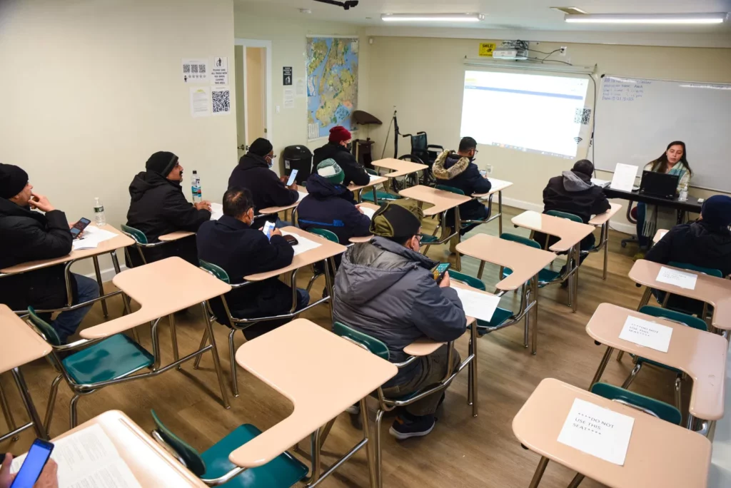 A classroom filled with students seated at desks, attentively facing the front where a teacher is giving a presentation. The students are dressed warmly, suggesting it’s a cold day, and some are using their phones while others have papers in front of them. The teacher is using a projector to display information on a screen. The classroom has a simple layout, with a map on the wall and signs on some desks indicating not to use certain seats, likely for social distancing purposes.