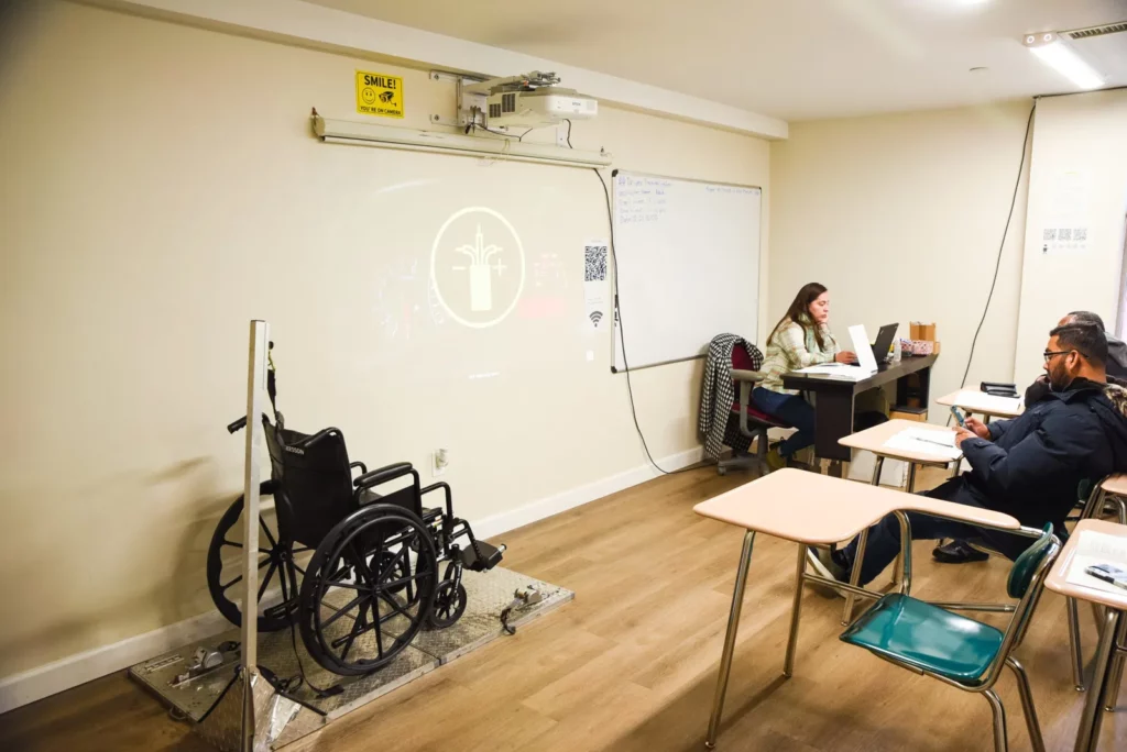 A classroom setting with a wheelchair positioned near the front, likely for a demonstration or training session. A projector displays an image on the wall, and a teacher is seated at a desk with a laptop, engaged in work. Two students sit at desks facing the instructor, one of them looking at their phone. The room has a simple, functional layout, with a whiteboard and a sign reminding people to ‘Smile! You’re on Camera.’