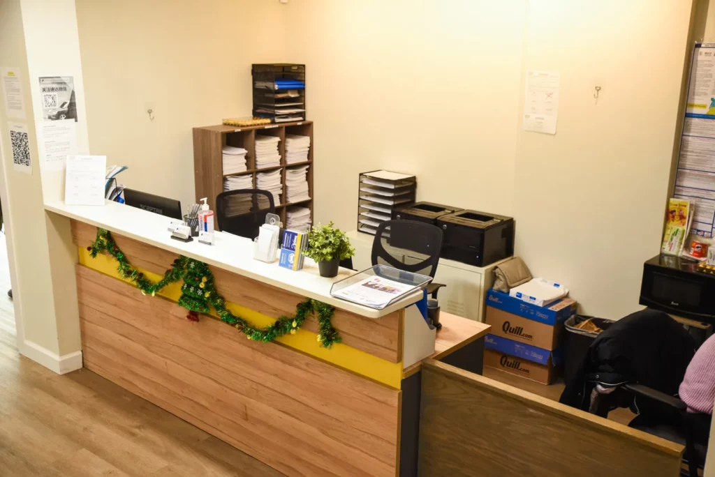 A tidy reception desk in an office setting, decorated with a festive green garland. The desk is equipped with office essentials, including a computer monitor, hand sanitizer, and paperwork. Behind the desk, there are shelves filled with neatly stacked papers and office supplies. The overall environment is organized and professional, with a touch of holiday cheer