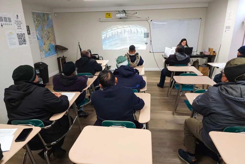 A classroom with students seated at individual desks, facing a projector screen at the front of the room where an instructor is teaching. The students are wearing winter clothing, indicating it may be cold outside. The projector displays an image related to the lesson, and there is a map on the wall. The environment is focused and conducive to learning, with students engaged in the material presented